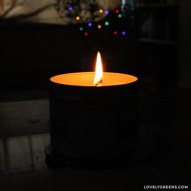 The silhouette of a candle flame coming out of a metal tin candle. It and colored Christmas lights are the only thing to be seen in a dark room.