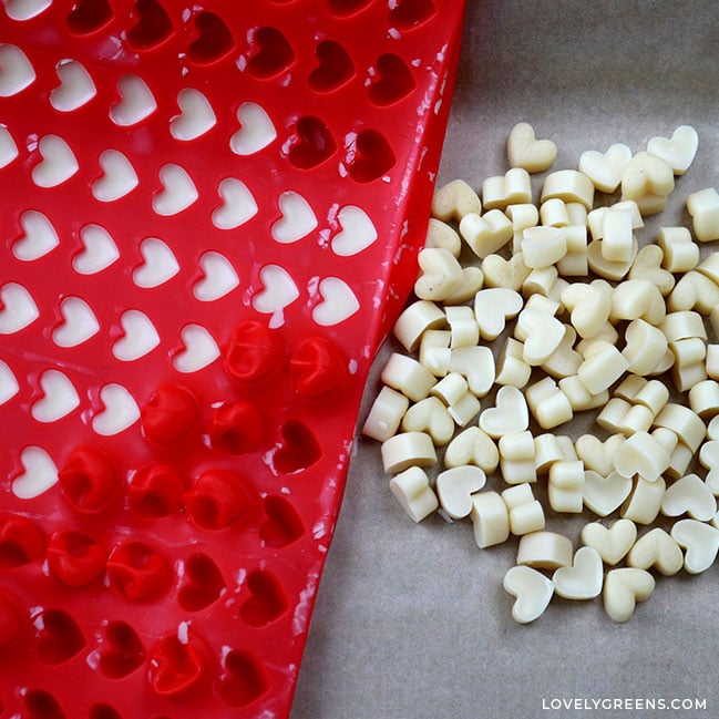 A pile of tiny heart-shaped lotion dots sits to the right of the red silicone mold they were popped out of.