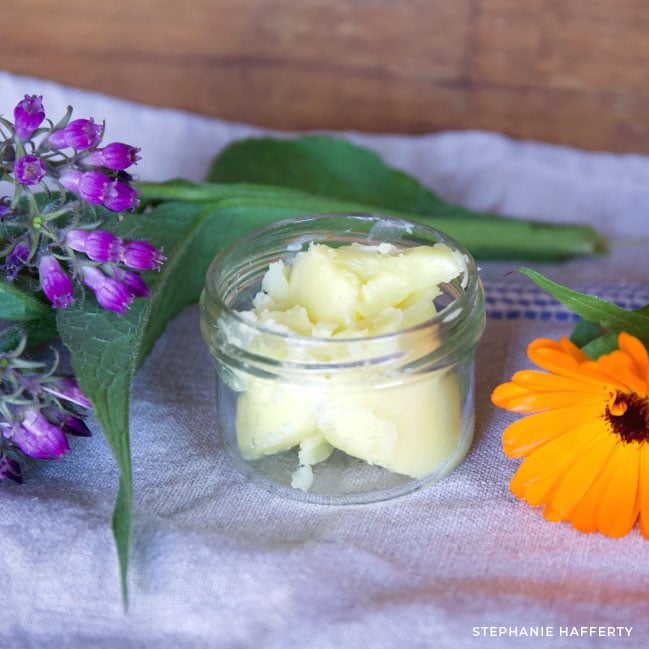 A small glass jar filled with scoops of salve sits on a white tablecloth. Fresh comfrey flowers and calendula flowers sit on either side.