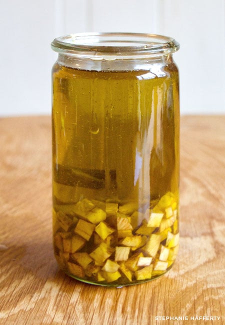 A tall glass jar filled with extra virgin olive oil and cubed comfrey root. The comfrey root is at the bottom and comes up to just under 1/3 of the jar.