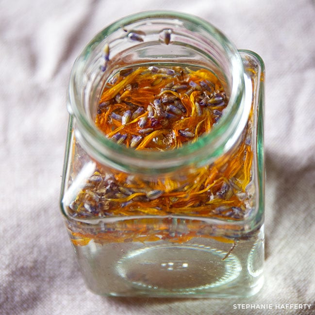 Looking down at a small open glass jar that's square with a short neck and round opening. Inside is what looks like clear water with a thin layer of calendula and lavender flowers floating on top.