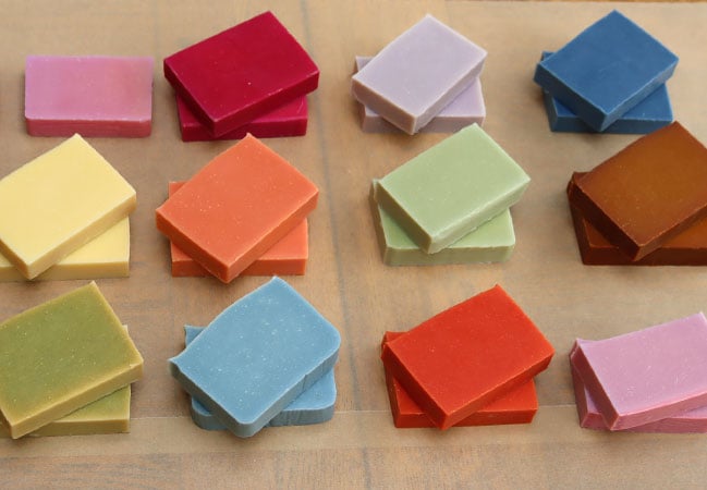 Twelve small piles of differently colored soap stacked on light brown paper.