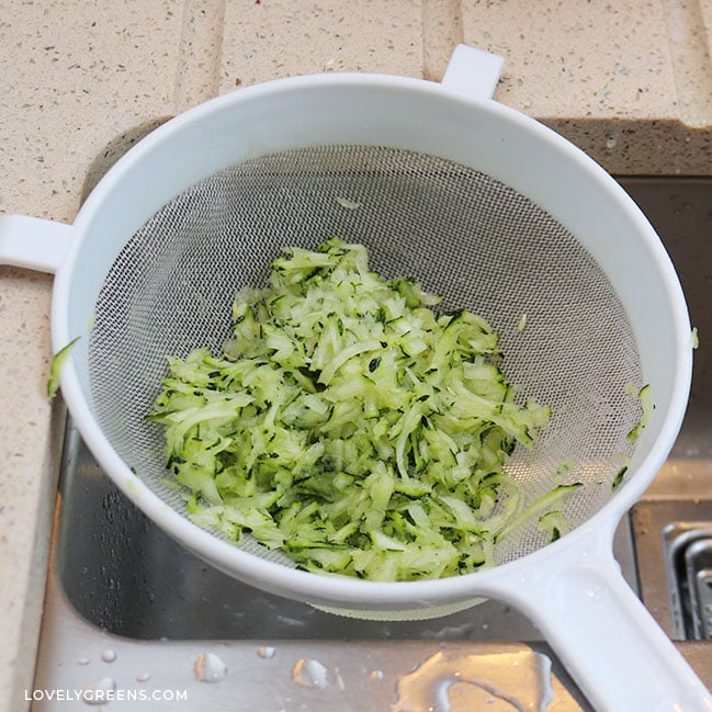 A cup of grated zucchini in a white sieve set over the small middle sink area in kitchen sink.