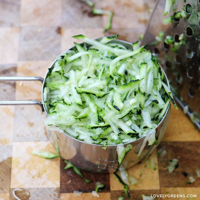 Close-up of a stainless steel measuring cup filled with freshly grated zucchini. It's pale green with dark green edges where the skin is visible.